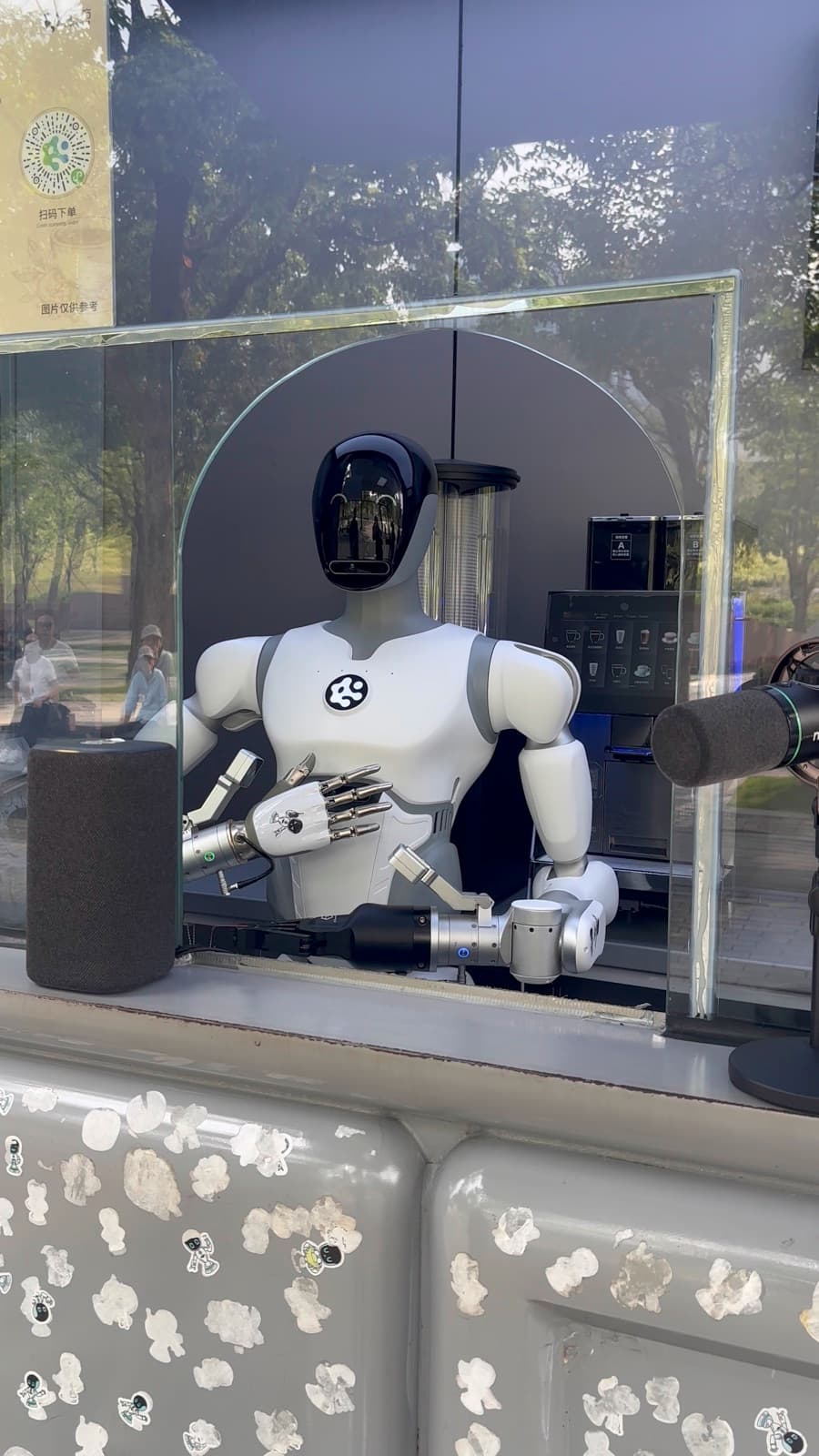 Humanoid robot behind a glass coffee counter near Shenzhen Bay Inner Lake, with trees reflected in the window.